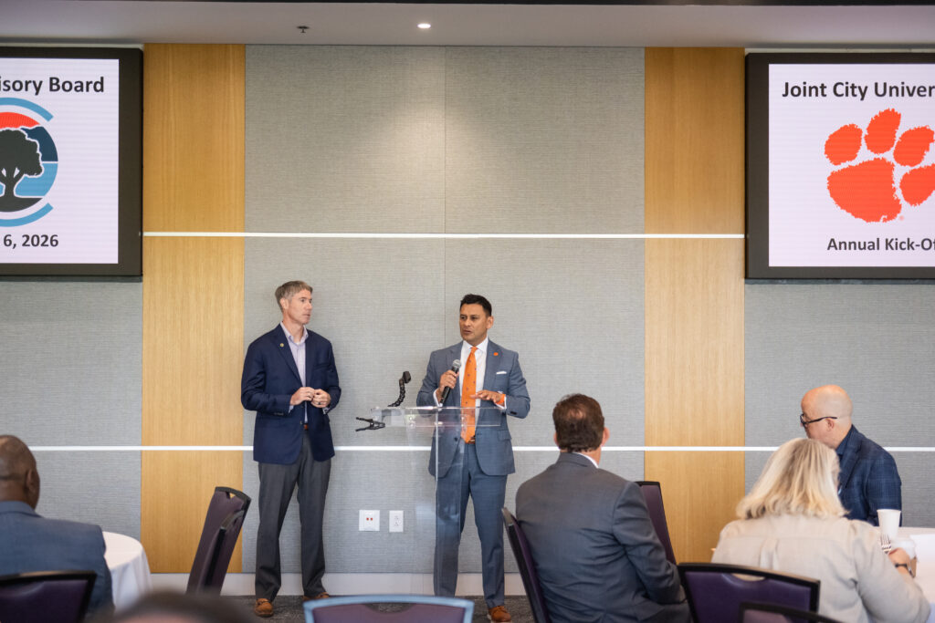 Two men in suits speak from a podium to an audience