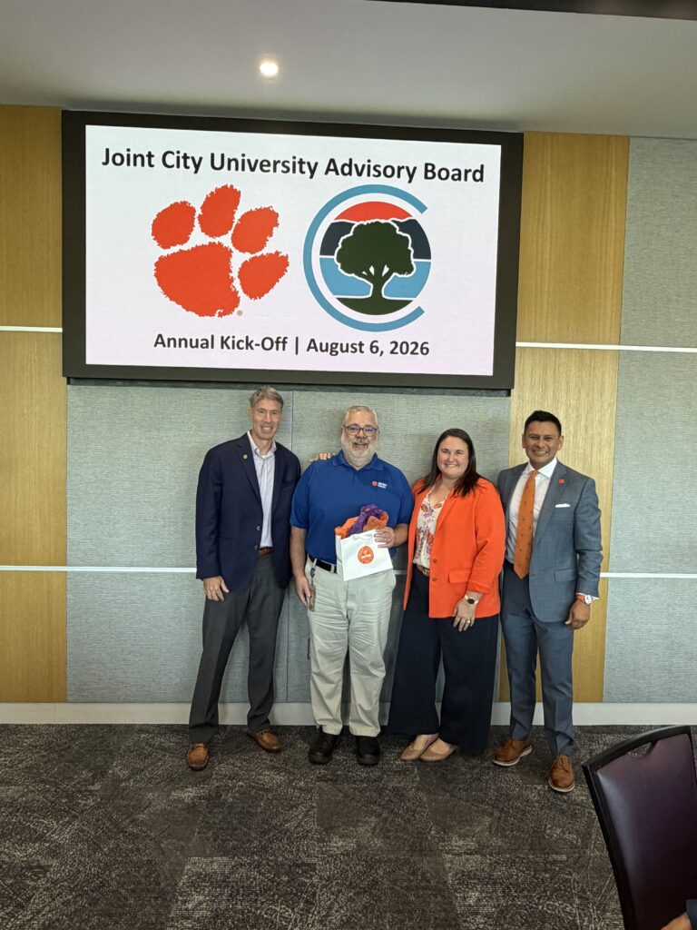 Four individuals in front of a Joint City University Advisory Board digital sign