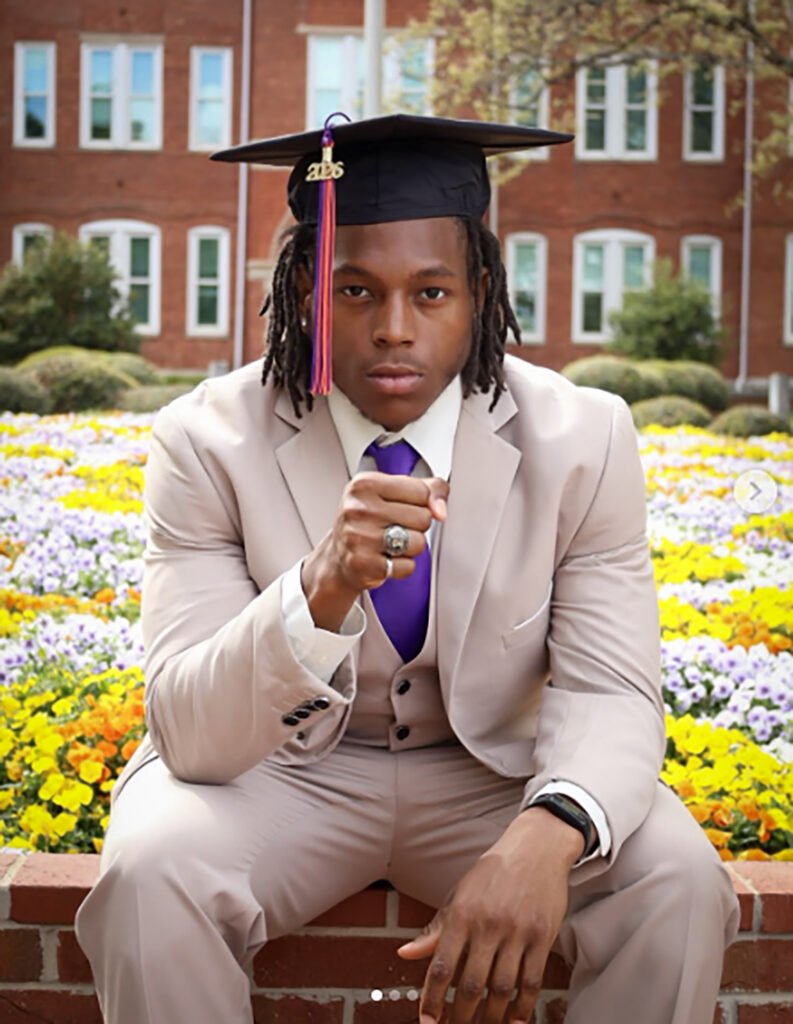An African American man wearing a tan suit and graduation cap sits on the edge of a flower bed, holding his hand with his class ring out to the camera