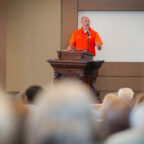 A man in an orange polo shirt speaks from behind a podium