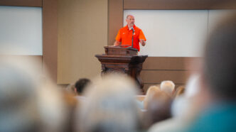 A man in an orange polo shirt speaks from behind a podium