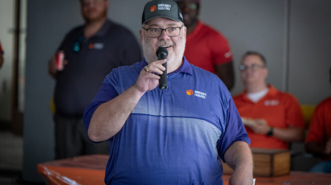 Todd Barnette, wearing a polo and hat, speaks into a microphone at an event.