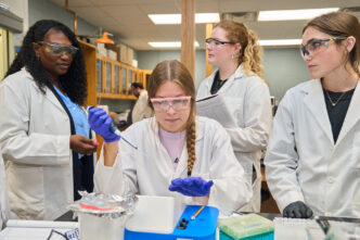 three students in lab coats talk with a Black woman wearing a lab coat in a lab at Clemson.