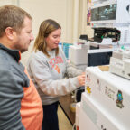 A man wearing a gray and orange sweatshirt watches a woman wearing a gray Clemson sweatshirt in a lab.