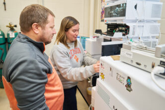 A man wearing a gray and orange sweatshirt watches a woman wearing a gray Clemson sweatshirt in a lab.