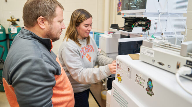 A man wearing a gray and orange sweatshirt watches a woman wearing a gray Clemson sweatshirt in a lab.