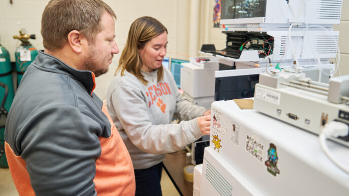 A man wearing a gray and orange sweatshirt watches a woman wearing a gray Clemson sweatshirt in a lab.