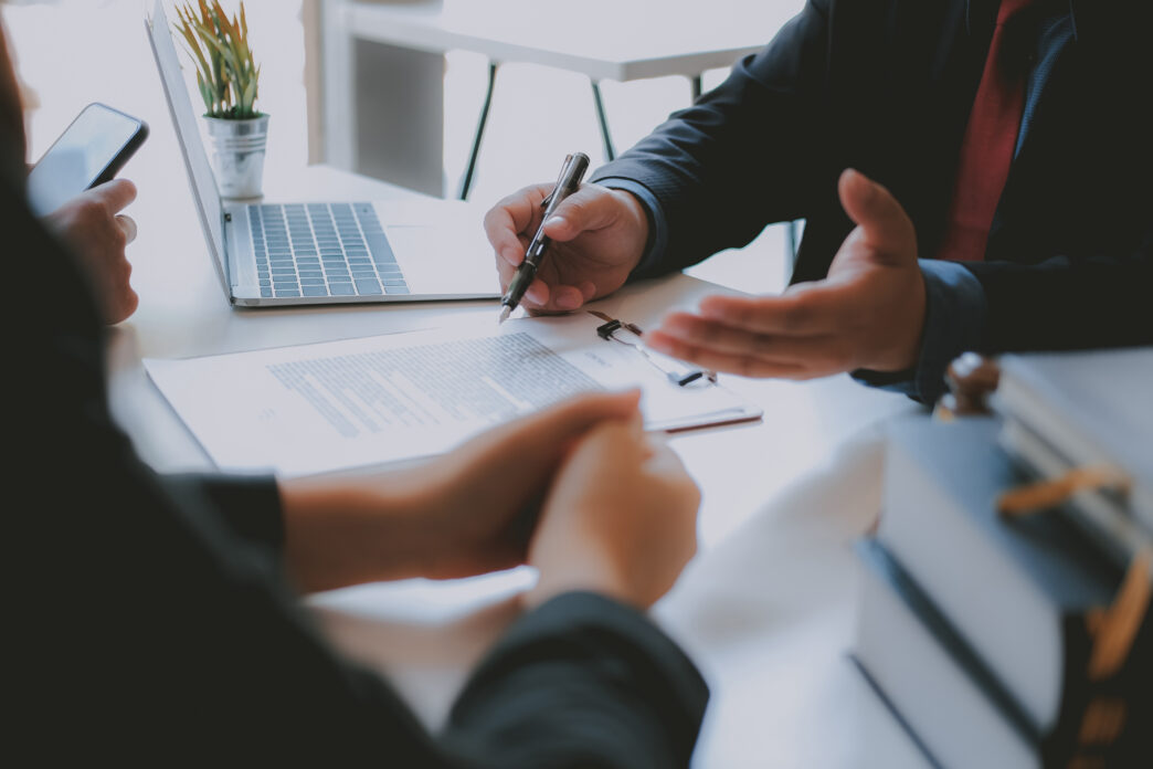 Two people's hands leaning on a desk. One person's hands are open and holding a pen, as if explaining while speaking.