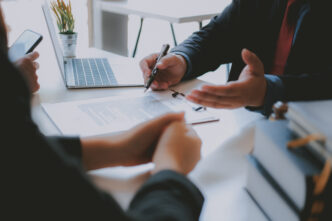 Two people's hands leaning on a desk. One person's hands are open and holding a pen, as if explaining while speaking.