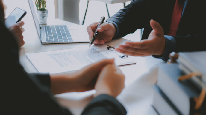 Two people's hands leaning on a desk. One person's hands are open and holding a pen, as if explaining while speaking.