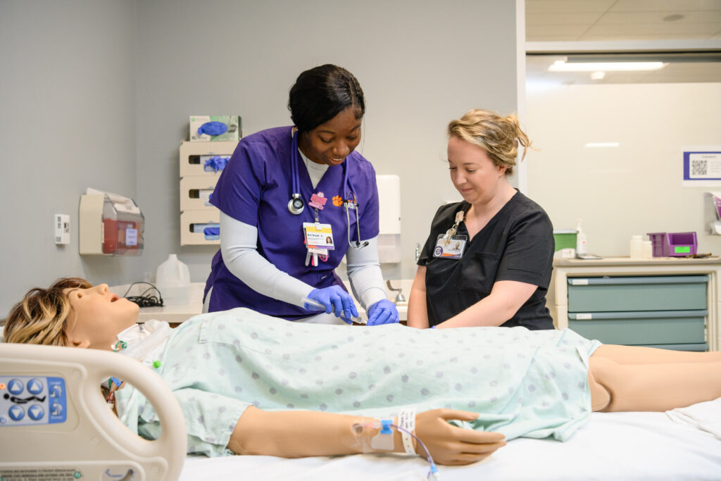 A young woman wearing purple nursing scrubs works alongside an older nurse wearing black scrubs in a hospital room. 
