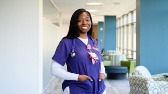 A young Black woman wearing a purple nursing scrub top with Clemson branding stands in a well-lit hallway.