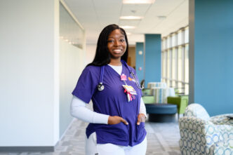 A young Black woman wearing a purple nursing scrub top with Clemson branding stands in a well-lit hallway.
