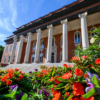 Low-angle view of Clemson University’s Sikes Hall framed by colorful orange and purple flowers under a bright blue sky.