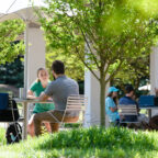 Students gather outdoors under umbrellas on Clemson’s campus, talking and spending time together at tables.