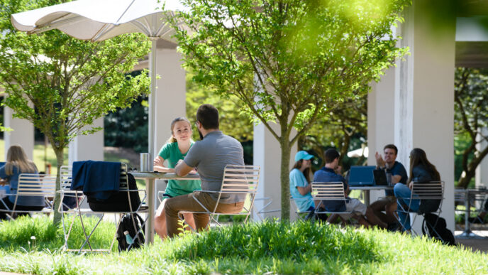 Students gather outdoors under umbrellas on Clemson’s campus, talking and spending time together at tables.