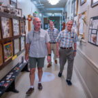 Three men walk down a hallway decorated with insect displays and posters.