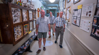 Three men walk down a hallway decorated with insect displays and posters.