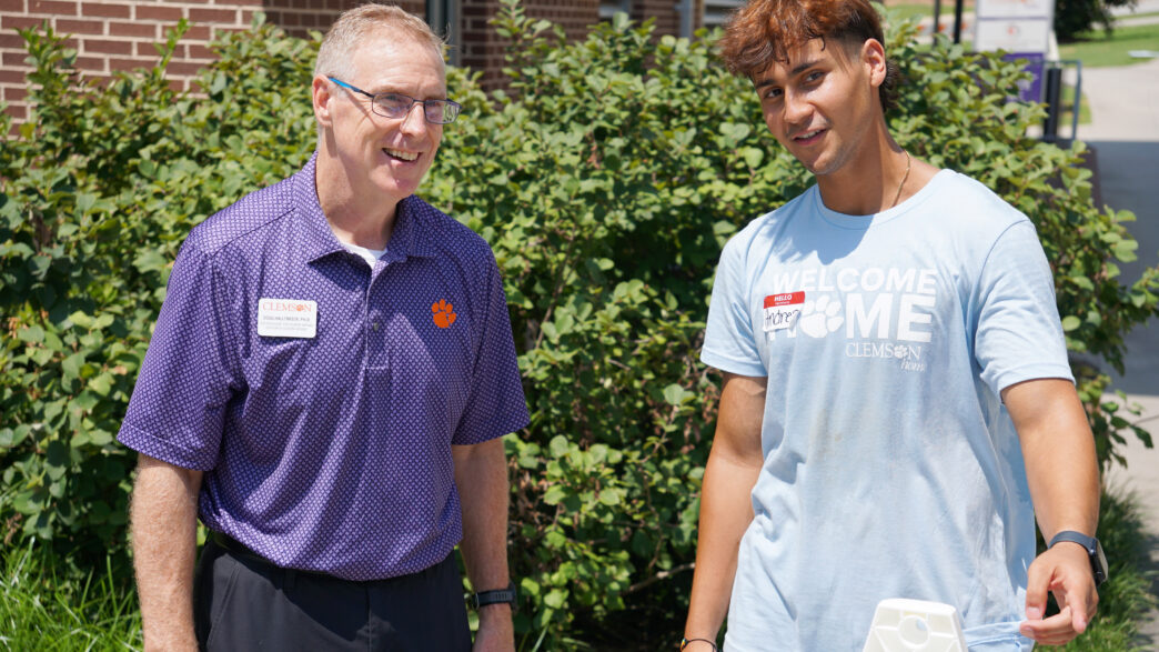 A male in glasses wearing a purple polo shirt speaks to a student wearing a Clemson Home t-shirt