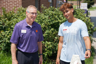 A male in glasses wearing a purple polo shirt speaks to a student wearing a Clemson Home t-shirt