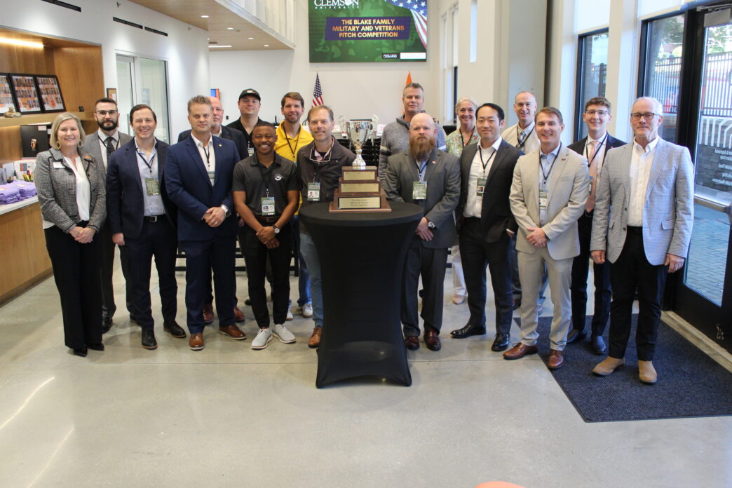 A group of 17 people pose standing around a table with a large trophy on it.