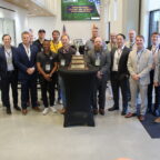 A group of 17 people pose standing around a table with a large trophy on it.