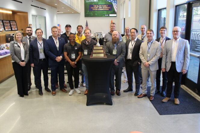 A group of 17 people pose standing around a table with a large trophy on it.