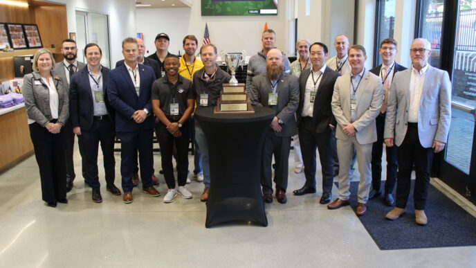 A group of 17 people pose standing around a table with a large trophy on it.