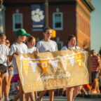 Students in white t-shirts hold a sign that reads "First Friday Parade 2025" while walking in front of a brick building