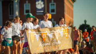 Students in white t-shirts hold a sign that reads "First Friday Parade 2025" while walking in front of a brick building