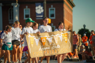 Students in white t-shirts hold a sign that reads "First Friday Parade 2025" while walking in front of a brick building