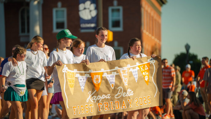 Students in white t-shirts hold a sign that reads "First Friday Parade 2025" while walking in front of a brick building