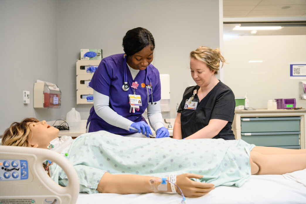 Two health care providers, one in purple scrubs and one in black scrubs, stand next to a hospital bed with a patient manikin lying in it