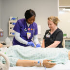 Two health care providers, one in purple scrubs and one in black scrubs, stand next to a hospital bed with a patient manikin lying in it