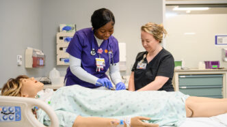 Two health care providers, one in purple scrubs and one in black scrubs, stand next to a hospital bed with a patient manikin lying in it