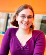 headshot of a woman with brown hair and glasses wearing a purple shirt with an orange wall in the background