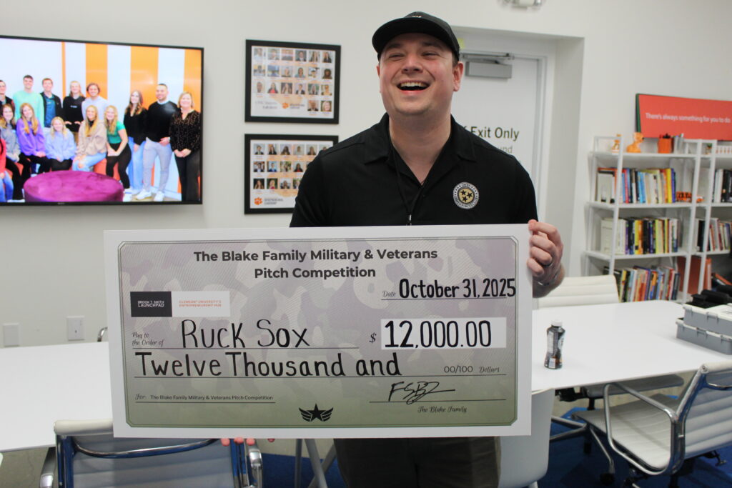 A man in a baseball cap and polo shirt holds a large check made out to Ruck Sox for $12,000, smiling broadly.