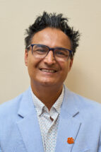 Headshot of a man wearing a blue suit jacket with an orange tiger paw pin and a collared shirt. He's got dark hair that sticks up and is wearing glasses (Shahid Mukhtar)
