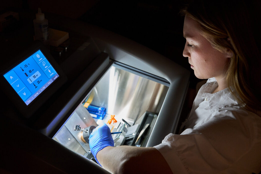 A woman wearing a white lab coat works with a machine that freezes and cuts tissue samples.