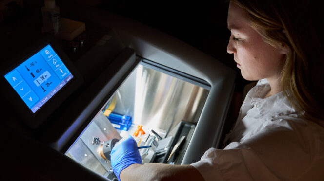 A woman wearing a white lab coat works with a machine that freezes and cuts tissue samples.