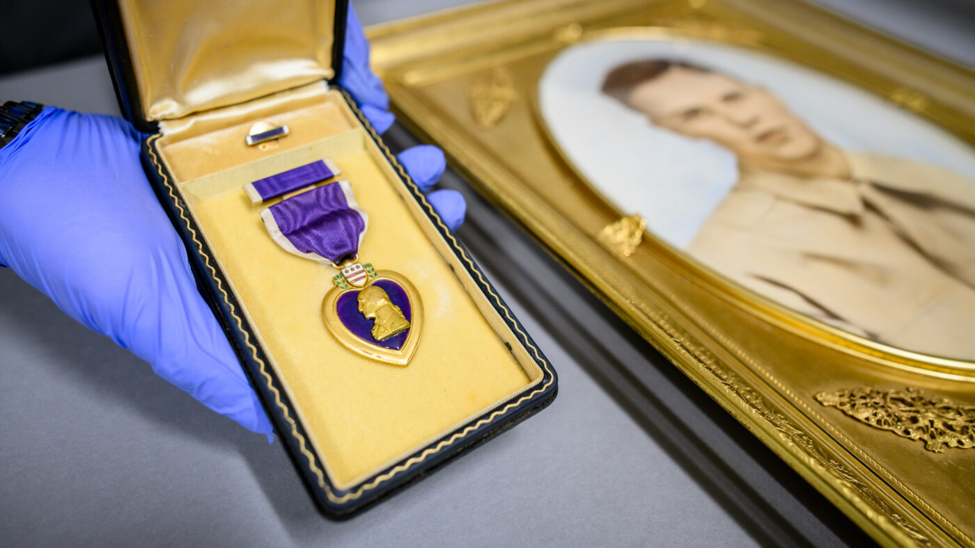 A gloved hand holds a small jewelry box containing a purple heart next to a golden-framed portrait of a young man in military uniform.