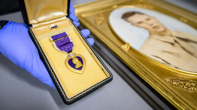 A gloved hand holds a small jewelry box containing a purple heart next to a golden-framed portrait of a young man in military uniform.