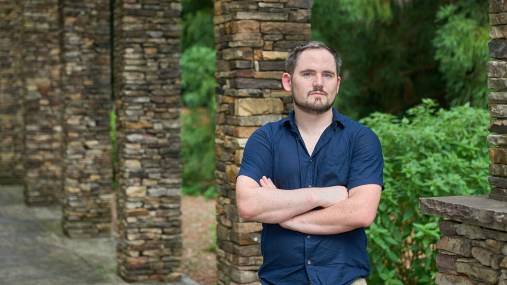 A man with dark hair and a beard and mustache wearing a dark blue button shirt stands with his arms crossed in front of a stone column with greenery in the background.