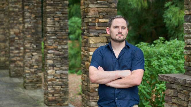 A man with dark hair and a beard and mustache wearing a dark blue button shirt stands with his arms crossed in front of a stone column with greenery in the background.