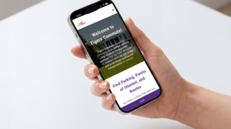 A hand holds an iPhone displaying the Tigers Commute app on its screen.