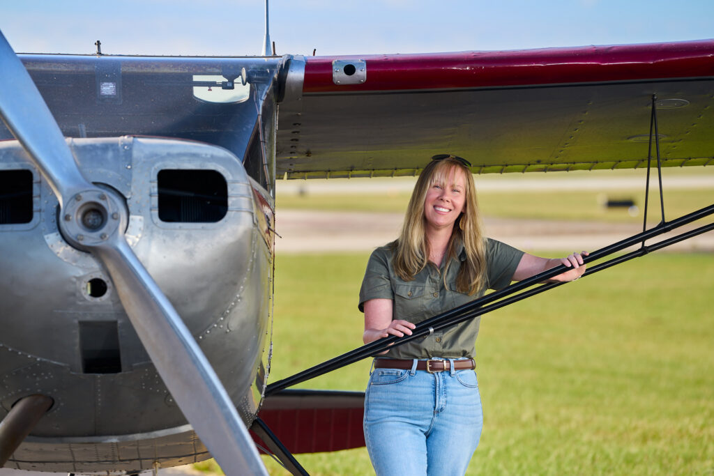 A woman wearing an olive green button shirt leans up against the strut on the wing of her single engine aircraft parked on the taxiway at an airport