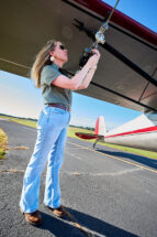 A woman wearing a green khaki shirt and jeans works on a device that is mounted on the wing of a plane and collects air samples.