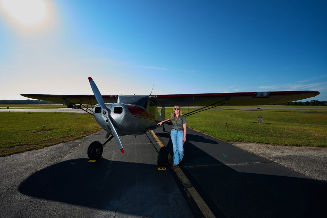 A woman wearing a blouse and blue jeans stands at the wing of a silver single engine aircraft on the runway