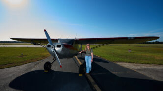 A woman wearing a blouse and blue jeans stands at the wing of a silver single engine aircraft on the runway