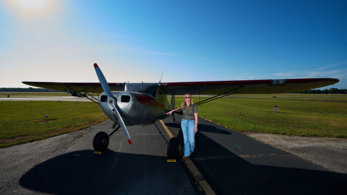 A woman wearing a blouse and blue jeans stands at the wing of a silver single engine aircraft on the runway